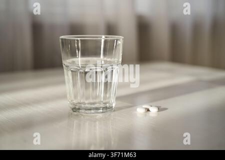 Glass Of Water With Two White Tablets Resting On Tabletop In Sunlit Interior Near Window With Sheer Curtains Soft Shadows And Reflective Surface On Pale Countertop. Stock Photo