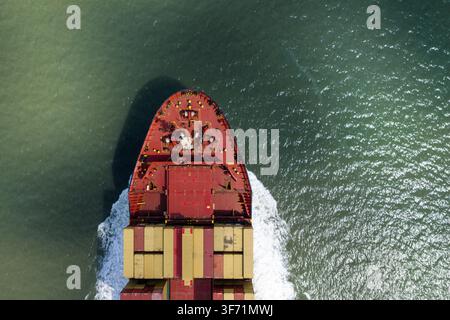 Aerial view of a large container ship cutting through the sea, with its red bow and stacked cargo containers visible as it moves through the water in Stock Photo