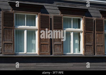 Vintage wooden house facade featuring double glass windows with rustic dark brown louvered shutters and dark painted timber cladding Stock Photo