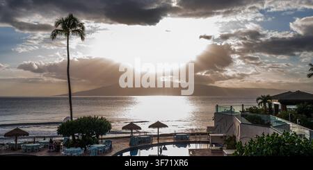 Sunbeams break through clouds over the ocean at dusk, viewed from a resort terrace with a palm tree and pool in the foreground. Stock Photo