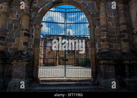 A locked wrought iron gate frames the view into the courtyard of crumbling stone colonial ruins beneath a bright blue sky. Stock Photo