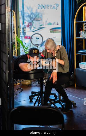 March 2023: A tattoo artist tattoos a client's arm near the open doorway of a studio, with reversed shop signage visible in the window. Stock Photo