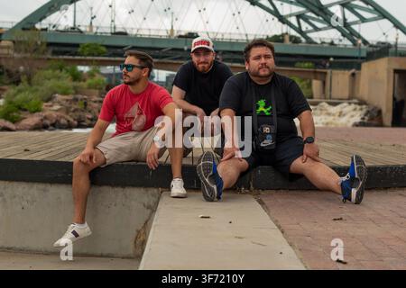 Riverside boardwalk - July 2022: Three men sit together on a concrete ledge along a riverside boardwalk, a large steel arch bridge spanning the water Stock Photo
