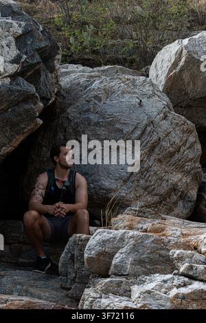 Southern Arizona - July 2022: A man sits in the shade of a rock crevice, surrounded by large weathered boulders, gazing off to the side. Stock Photo
