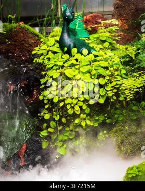 A small green dragon figurine sits among moss and trailing plants beside a misty miniature waterfall. Stock Photo