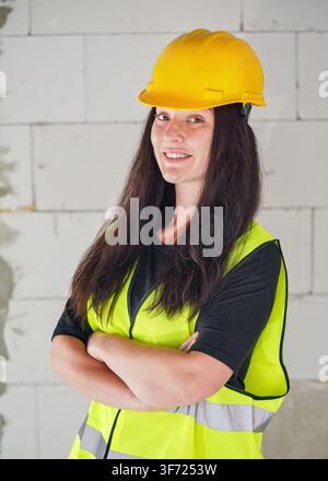 Young woman in yellow hard hat and green high visibility vest, long dark hair, hands crossed, looking into camera. Blurred construction site wall back Stock Photo