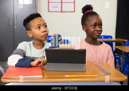 African American female pupils child, teen sitting at classroom desk wearing sweaters using tablet. Students, learning, school, collaboration, device, Stock Photo