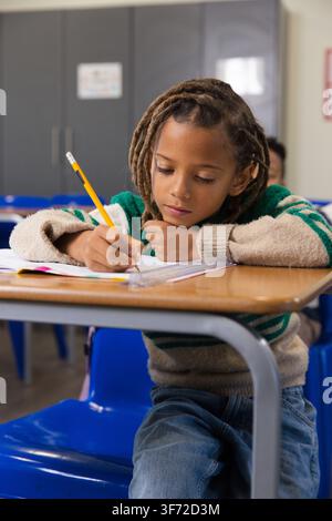 African American male student writing in notebook at school desk holding yellow pencil in sweater. Classroom, education, study, learning, focus, conce Stock Photo