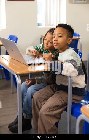 African American students sitting at wooden desk using silver laptop, wearing striped sweaters. Collaboration, classroom, daylight, computer, youth, p Stock Photo