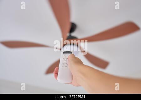 Hand holding remote control aimed at ceiling fan in home interior. Concept of home cooling, climate control and indoor comfort Stock Photo