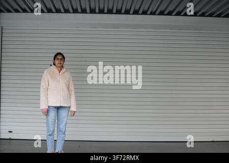 Woman stands in front of large white garage door. She wears light-colored jacket and blue jeans. Her posture is relaxed and upright. Background is pla Stock Photo