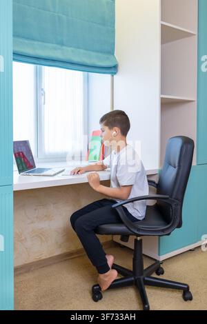 focused boy using laptop in home office side view with chair and desk, white and black background, natural light Stock Photo