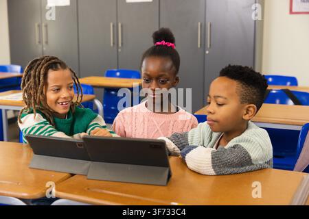 School-age classmates leaning around tablet in folio case at wooden desks, pointing. Classroom, collaboration, learning, group, education, device, foc Stock Photo