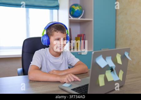 focused boy using laptop in home office with headphones and wooden desk, blue and white background Stock Photo