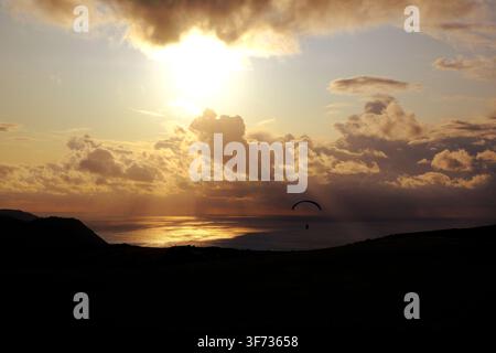 Contre-jour silhouette of a paragliderin under a stunning sunset panorama, golden light reflecting on the sea, UK Stock Photo