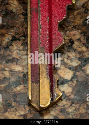 Extreme close up macro shot of the serrated blade of a weathered brass door key with worn red paint resting on a granite stone surface Stock Photo