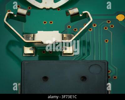 Extreme close up macro shot of electronic components including a metal battery contact clip and a black microchip mounted on a green printed circuit b Stock Photo
