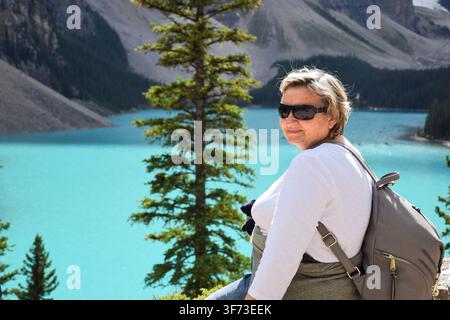 Side view of senior woman with backpack and sunglasses posing in front of turquoise mountain lake with evergreen tree and rocky mountain background Stock Photo