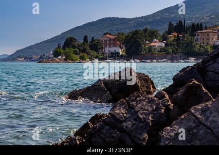 Large dark rocks sit along a coastal shoreline with calm blue water in front of boats near a small harbor. Buildings and trees. Stock Photo