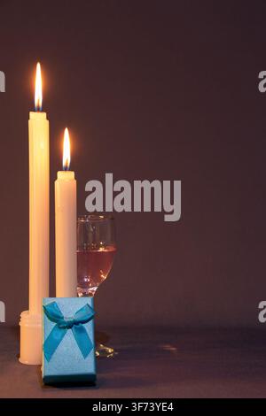 A still life of two lighted candles, a glass of wine, a white glass holder and a blue-ribboned gift box, against a gradient pink backdrop. Stock Photo
