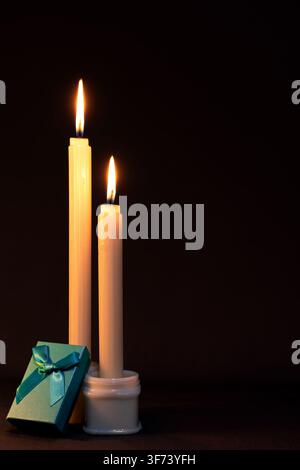 Two tall candles alight, with a blue gift box propped against a white bottle being used as a candle holder, as if presenting a gift. Dark backdrop. Stock Photo