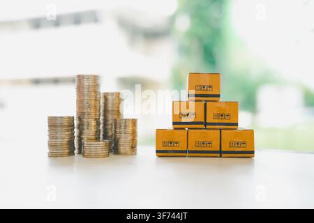 Stacked coins beside cardboard parcel boxes on white background, symbolizing shipping costs, e-commerce investment, logistics budgeting, and financial Stock Photo