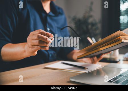 Professional holding eyeglasses while reading an open book beside a laptop, representing adult education, self-study, digital eye strain, reading fati Stock Photo
