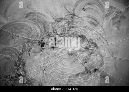 Monotone view of spiderweb cracks radiating across a smeared glass window pane from a central impact point Stock Photo