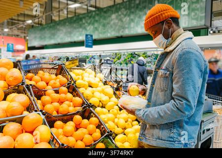Young black man wearing a protective face mask while selecting fresh lemons and oranges in the produce section of a grocery store, making health conscious choices Stock Photo
