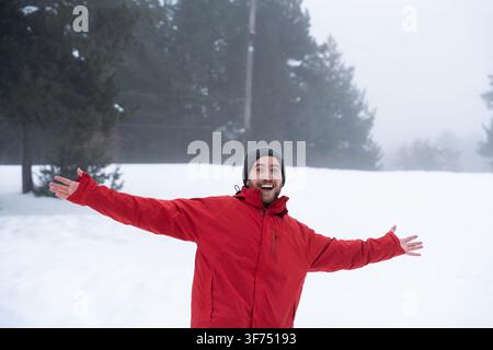 Enthusiastic man wearing red winter jacket and grey beanie hat standing outdoors in snowy forest with arms open expressing freedom and joy in the fog Stock Photo