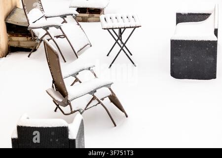 Outdoor furniture covered in fresh white winter snow on a patio surface with multiple chairs and a small table arranged outdoors during a cold season Stock Photo