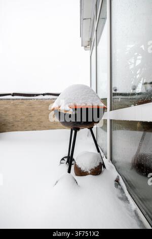Round charcoal grill covered in fresh white snow sits on a residential terrace beside a large glass wall, a stone fence in the background during a col Stock Photo