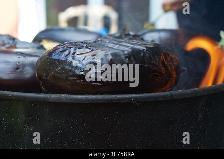 Close-up of fresh whole eggplants grilling on a hot barbecue pit with open flames and smoke rising from the glowing charcoal underneath. Stock Photo