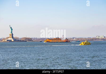 New York, USA - February 20, 2026: Statue of Liberty and iconic orange Staten Island Ferry sailing with a yellow water taxi on New York Harbor Stock Photo