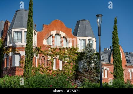 Elegant red brick residential building with ivy covered facade and tall cypress trees under blue sky Stock Photo
