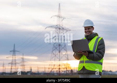 Electrical engineer in a safety vest smiles while using a laptop next to power lines at sunset. The sky shows colors of sunset in the background. Stock Photo