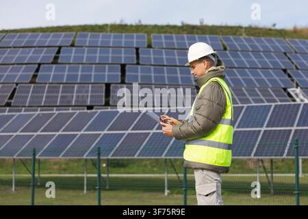 A worker stands in front of solar panels while using a tablet. The site is green with multiple rows of panels. Stock Photo