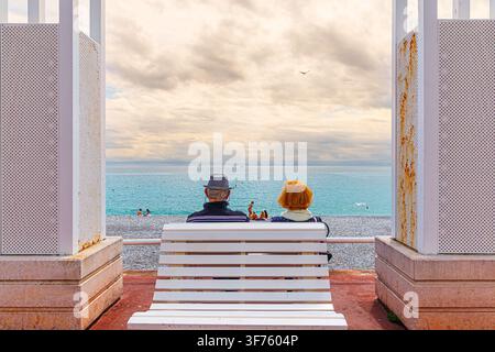 Elderly couple sits on a bench beneath a pergola on the Promenade des Anglais, looking over the sea from a pebble beach in Nice, France. Stock Photo