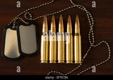 Two metal dog tags with chain lie beside five rifle cartridges on a dark wooden surface. Stock Photo
