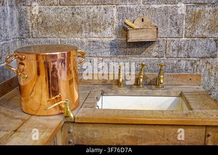 Polished copper hot water urn with a brass tap rests beside a wooden-rimmed scullery sink featuring three antique brass water faucets at Castle Drogo, Stock Photo