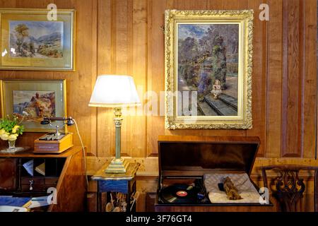 An antique wind-up phonograph record player sits open next to an electric table lamp, a candlestick telephone intercom, and framed oil paintings on a Stock Photo