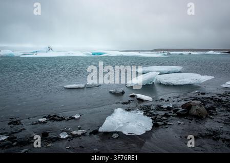 Small and large fragments of transparent and blue ice rest on the wet shore of dark sand and pebbles. In the background, the rippled water holds massi Stock Photo