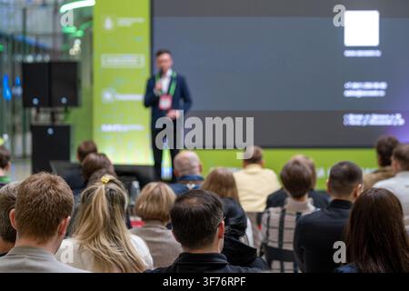Speaker addresses a seated audience beside a dark presentation slide on a large screen. Stock Photo