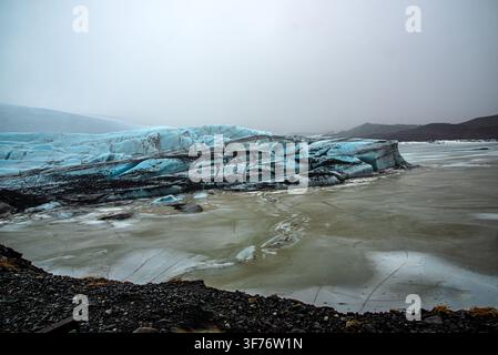 Wide view of the glacier in Iceland. In the foreground is a dark pebble shore, followed by a frozen and muddy lagoon. In the center rises the massive Stock Photo