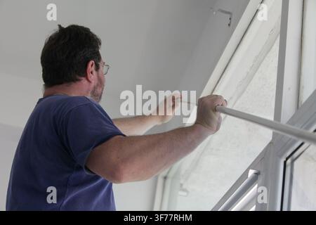 Man positioning a long curtain rail beside a window as part of a home curtain installation, preparing to mount the rail in place for window covering. Stock Photo