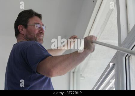 Man positioning a long curtain rail beside a window as part of a home curtain installation, preparing to mount the rail in place for window covering. Stock Photo