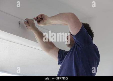 Man installing curtain rod support brackets with a screwdriver above a window during home improvement work. Practical DIY installation, home repair, r Stock Photo