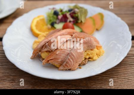 Juicy smoked salmon on scrambled eggs and bread with a colorful salad garnish on a white plate and a rustic wooden table, selected focus, narrow depth Stock Photo