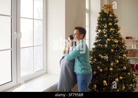 On Christmas morning, couple is standing together by large window wrapped in plaid. Stock Photo
