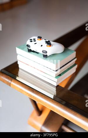 White Google Stadia controller resting atop a stack of books on a wooden and glass side table in a Stock Photo